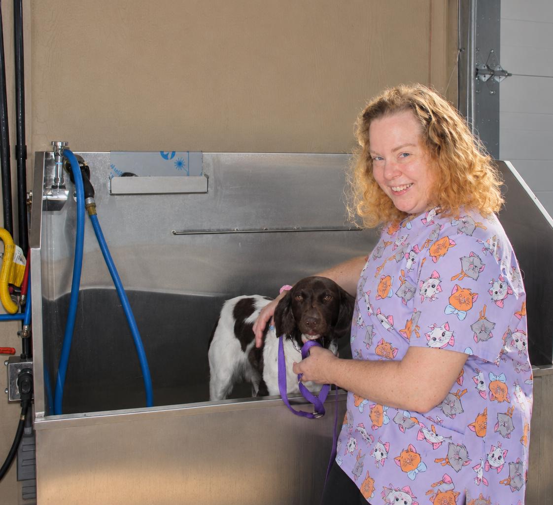 Angela smiling in a client's garage, one hand resting on a dog standing in a stainless wash station